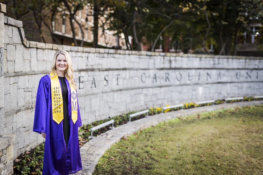 photo of a student in graduation regalia standing next to the large stone east carolina university sign on campus