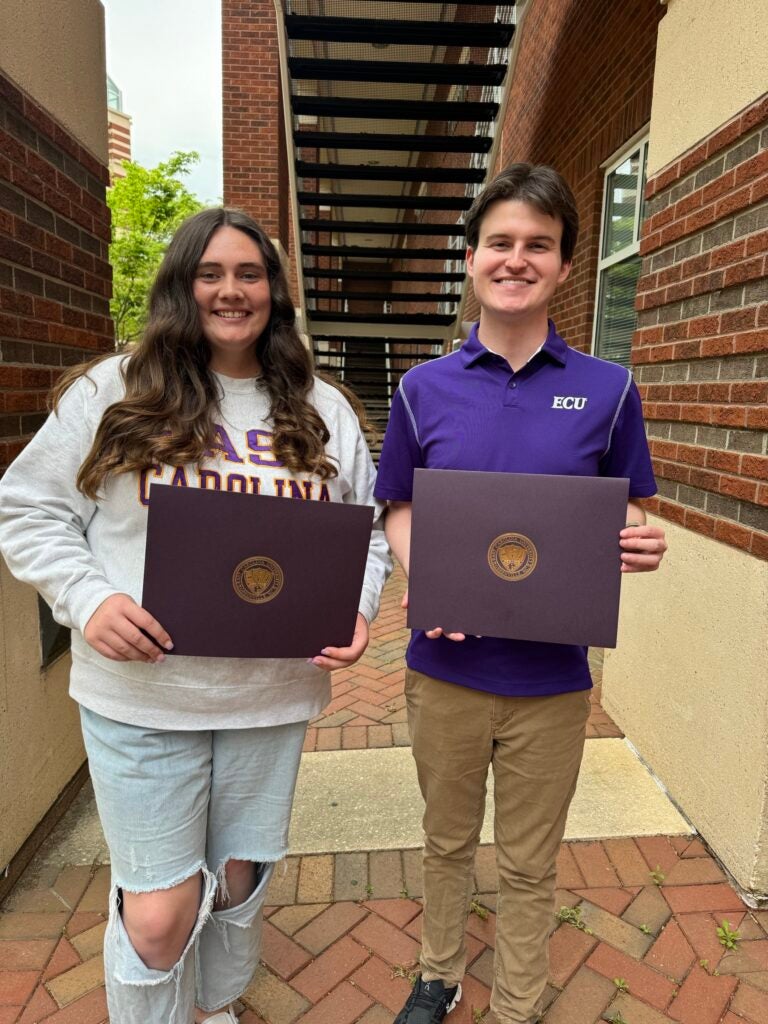 Christina Dowd is wearing a white sweat shirt with light blue jeans and Sam Huffman is wearing a purple shirt with brown pants. They are posing with their ECU Student Employee of the Year Certificate. The background is stairwell with a brick floor.