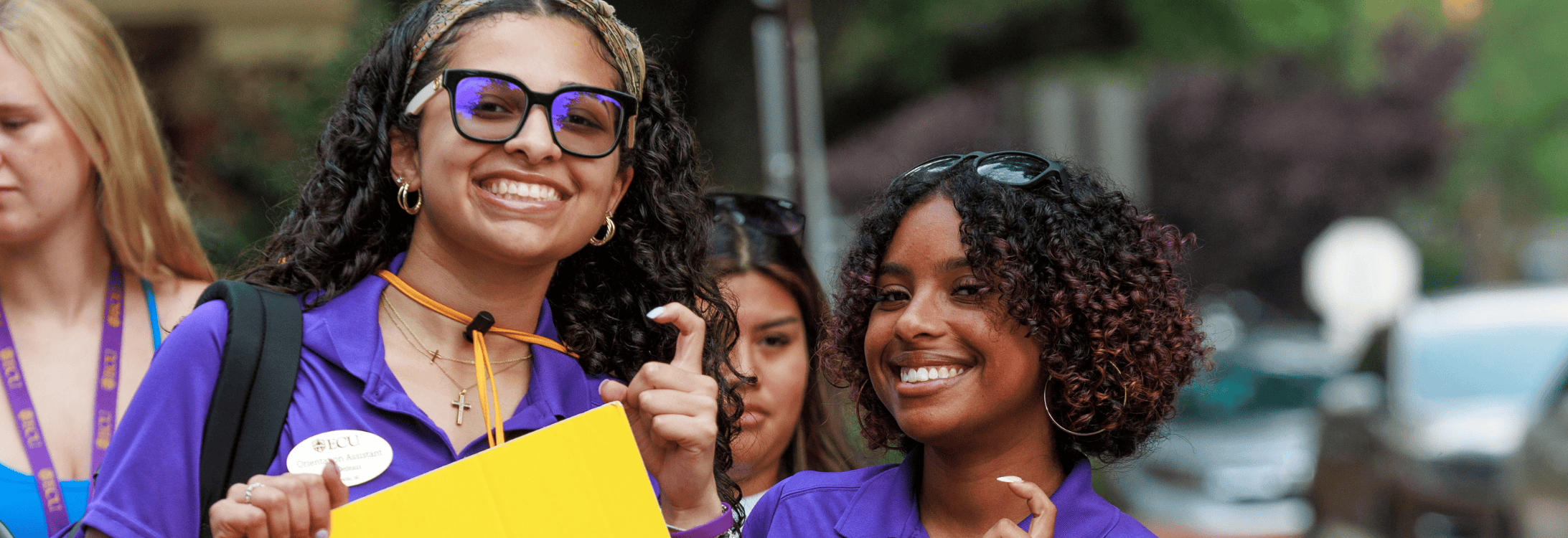 Two Orientation Assistants standing on campus, both smiling at the camera with students in background.